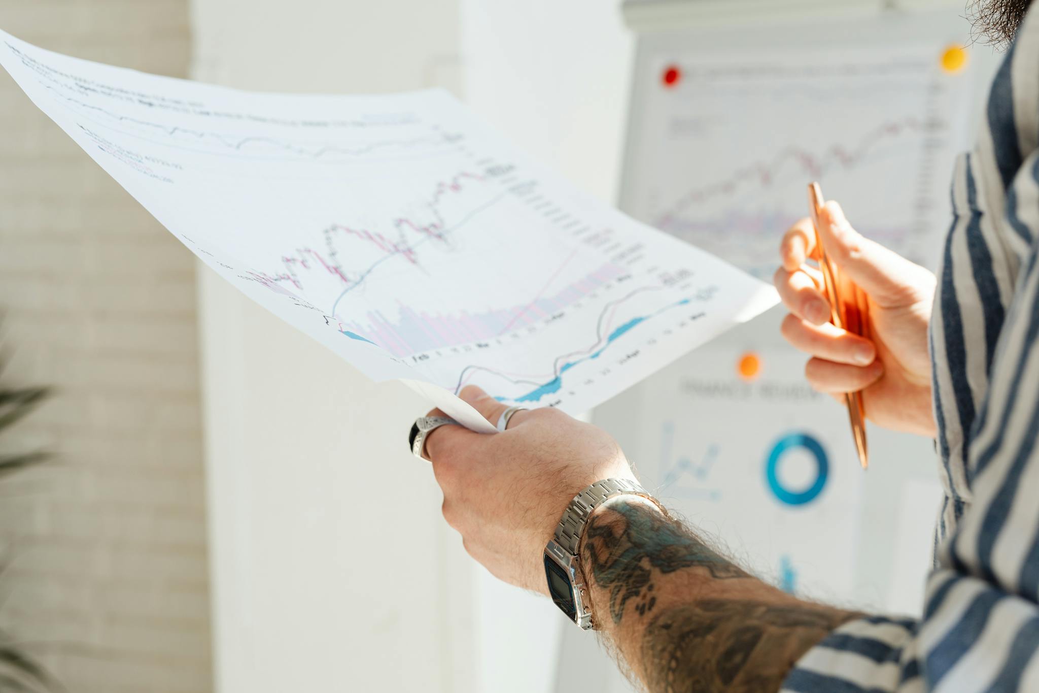 Close-up of a tattooed man's hands holding financial charts, with focus on data analysis and stock market trends.